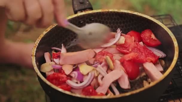 잘게 썬 소시지와 버섯과 토마토와 양파를 섞은 한 여인이 숯불 위에 프라이팬을 놓고 있다. — 비디오