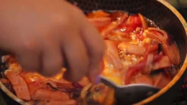 Жіноча рука змішує яєчня з овочами та ковбасою на сковороді на вугіллі з ложкою . — стокове відео