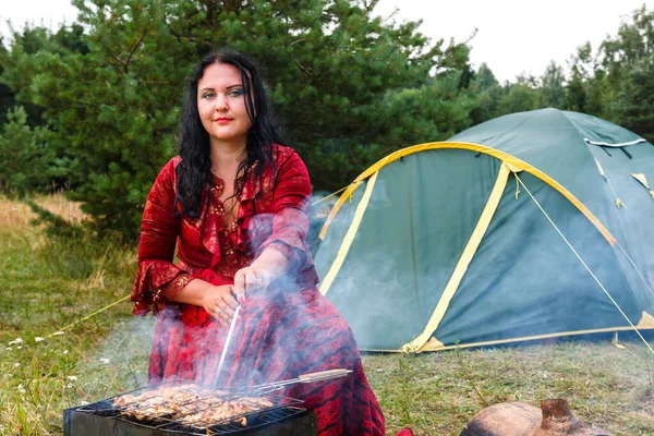 Молодая умная цыганка жарит мясо на гриле возле палатки в дыму от огня.. — стоковое фото