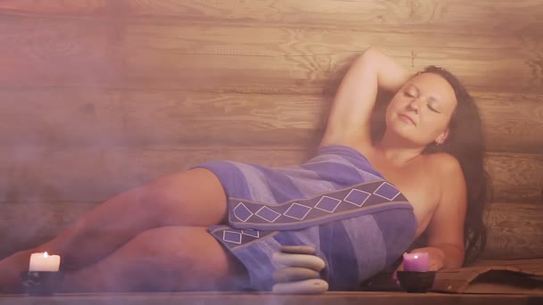 Uma mulher morena em uma sauna com velas está em uma toalha de relaxamento . — Vídeo de Stock