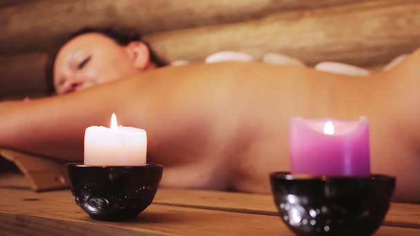 Une femme brune dans un salon de spa se couche avec des pierres sur son massage du dos — Video