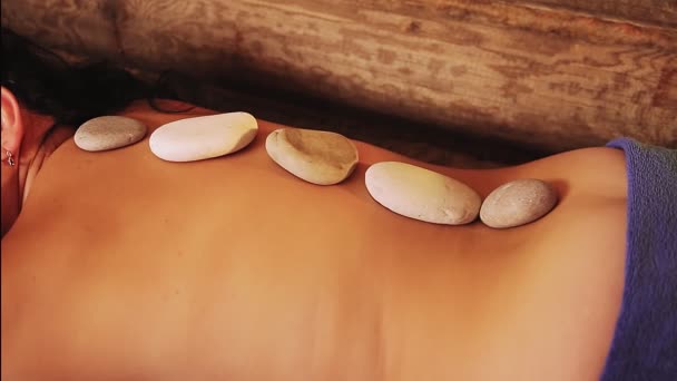 Een brunette vrouw in een spa salon ligt met stenen op haar rug met stenen therapie — Stockvideo
