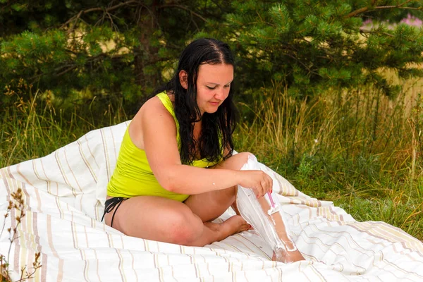 A young woman sits on a bedspread in a clearing shaves her legs with a razor. — Stock Photo, Image