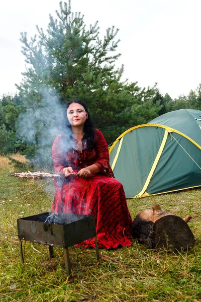 Молодая цыганка жарит мясо на гриле возле палатки в дыму от огня.. — стоковое фото