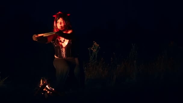 Una mujer en forma de demonio de negro con cuernos por la noche junto al fuego con un cuchillo antes del sacrificio — Vídeos de Stock
