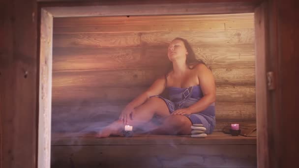 Een brunette vrouw in een sauna met kaarsen zit in een ontspannende handdoek — Stockvideo