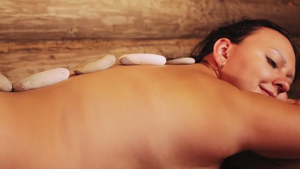 Uma mulher morena em um salão de spa está com pedras nas costas para uma massagem de relaxamento — Vídeo de Stock