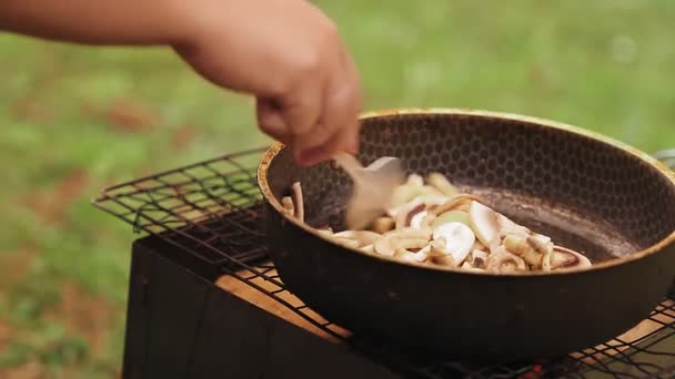 Uma mulher está mexendo cogumelos fritos com uma colher em uma frigideira nas brasas. — Vídeo de Stock
