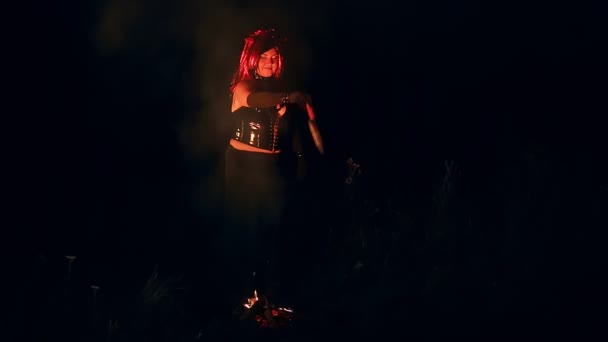 Une femme à l'image d'un diable en noir avec des cornes la nuit près du feu avec un couteau danse une danse rituelle. — Video