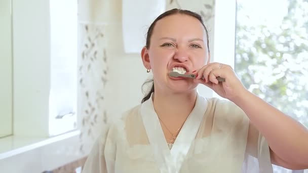 Una joven morena con una bata limpia los dientes en el baño por la mañana — Vídeos de Stock