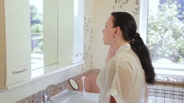 Una joven morena con albornoz delante de un espejo limpia los dientes en el baño por la mañana — Vídeo de stock