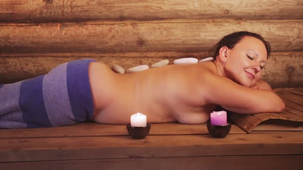 Брюнетка в спа лежит с камнями на спине массаж — стоковое видео