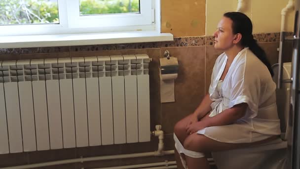 Een vrouw in een wit gewaad zit op het toilet in het toilet. — Stockvideo