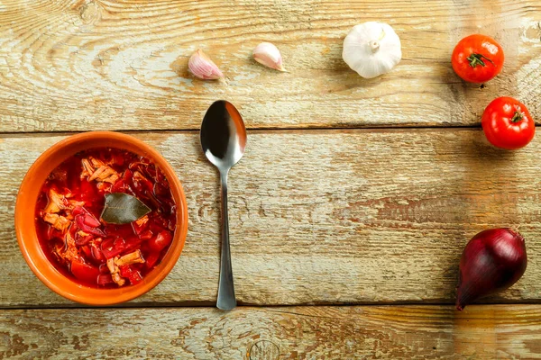 Um prato de borscht de barro com frango com uma colher em uma mesa de madeira perto de verduras. — Fotografia de Stock