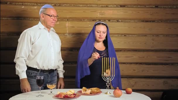 Joodse familie man en vrouw naast de feesttafel een vrouw steekt kaarsen aan een man houdt een glas voor kiddush. — Stockvideo