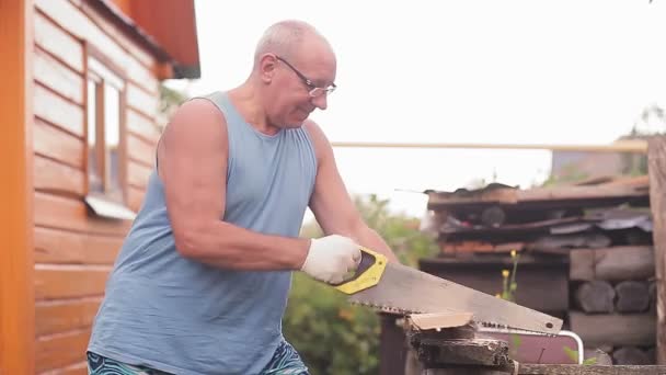 Un hombre en el patio sostiene una sierra de mano y la usa para cortar tablas. — Vídeo de stock