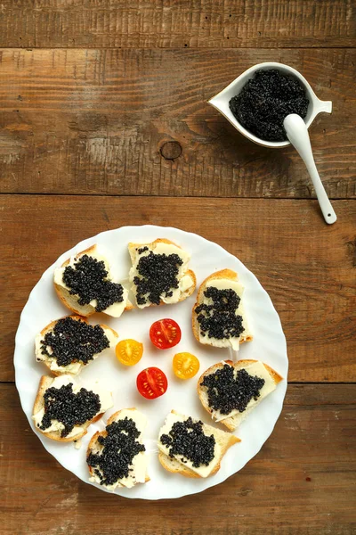 Sanduíches com caviar preto em uma placa branca em uma mesa de madeira ao lado de um prato de caviar com uma colher. — Fotografia de Stock
