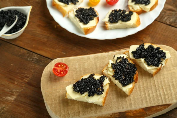 Sanduíches com caviar preto em uma tábua de madeira em uma mesa de madeira ao lado de um prato de caviar. — Fotografia de Stock