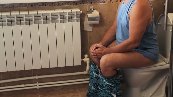 A man in shorts in the toilet sits on the toilet with a paper shot without a face — Stock Video