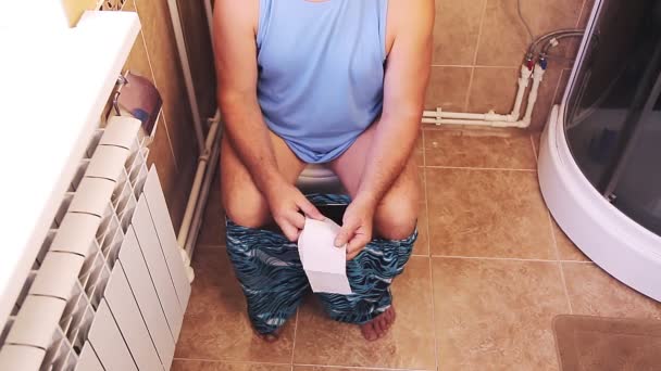 Um homem de calções e óculos no banheiro senta-se no banheiro sem rosto — Vídeo de Stock