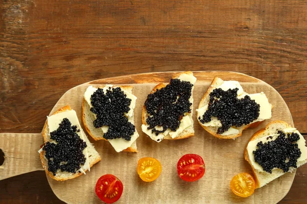 Sanduíches com caviar preto em uma placa de madeira em uma mesa de madeira ao lado de um prato de caviar com uma colher. — Fotografia de Stock