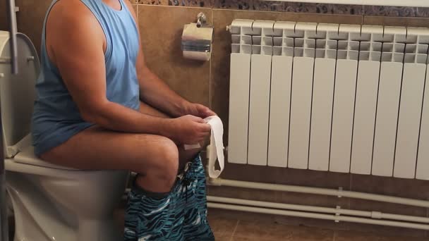 A man in shorts in the toilet sits on the toilet with a paper shot without a face. — Stock Video