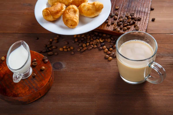 Кружка кофе со сливками и кувшин сливок и круассанов на столе среди разбросанных кофейных зерен. — стоковое фото