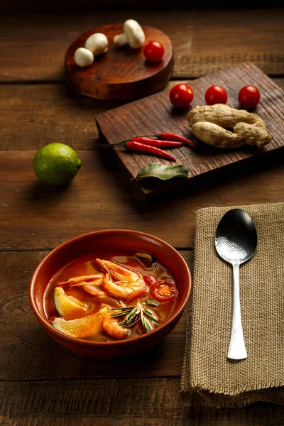 Sopa de ñame Tom con camarones y lima en un plato marrón en una mesa de madera junto a una cuchara en una servilleta e ingredientes jengibre de lima. —  Fotos de Stock