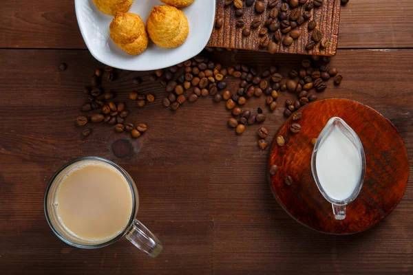 Кружка с кофе с молоком и кувшин сливок и круассанов на столе среди разбросанных кофейных зерен. — стоковое фото