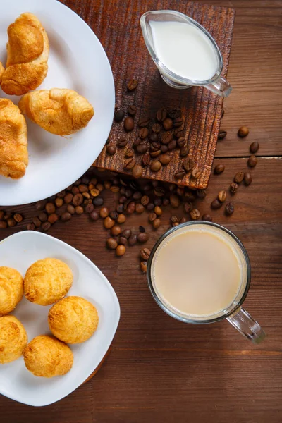 Şeffaf bir kupa, kremalı kahve, bir sürahi krema, kruvasan ve kahve çekirdekleri arasında masanın üstünde kurabiyeler..