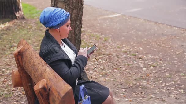 Mujer judía en un tocado tradicional en un banco en un parque de otoño escucha música con auriculares. — Vídeos de Stock