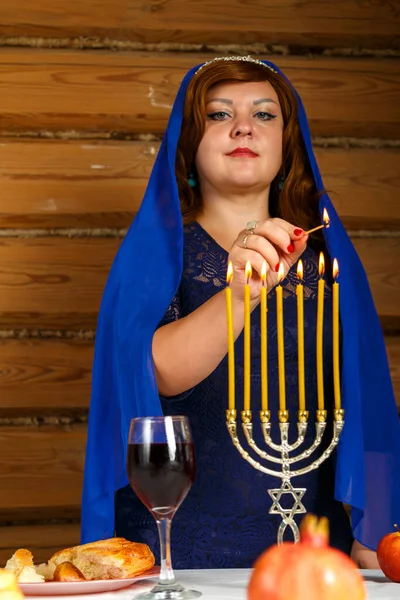 Een Joodse vrouw in een blauwe cape op haar hoofd steekt kaarsen aan op de avond van Rosh Hashanah in een menorah-vormige kandelaar. — Stockfoto