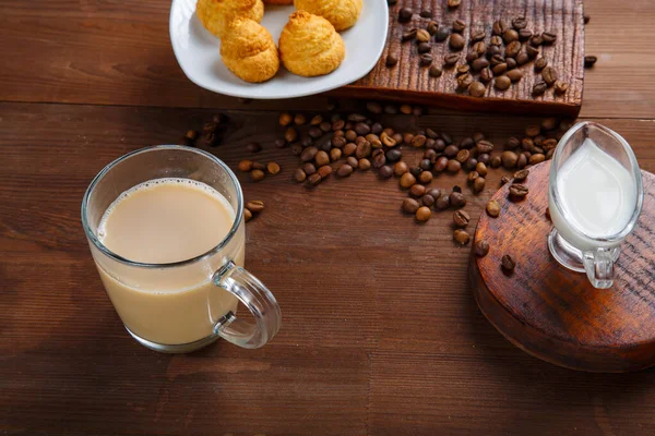 Кружка кофе со сливками и кувшин сливок и круассанов на столе среди разбросанных кофейных зерен. — стоковое фото