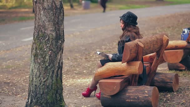 Eine Frau mit braunen Haaren im Hutprofil in roten Stiefeln und roter Tasche liest im Herbst auf einer Parkbank ein Buch. — Stockvideo