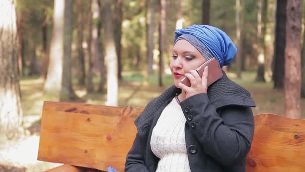 Una mujer judía en un tocado tradicional de perfil en un banco en un parque de otoño habla por teléfono. — Vídeos de Stock