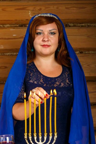 Una bella donna ebrea con un mantello blu sulla testa accende candele alla vigilia di Rosh Hashanah in un candeliere a forma di menorah. — Foto Stock