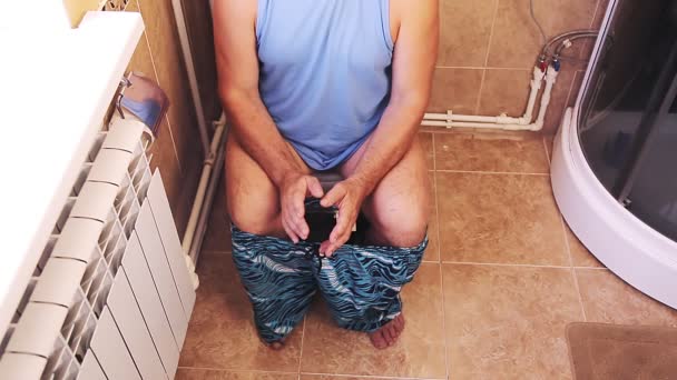 Een man in korte broek en bril in het toilet zit op het toilet zonder gezicht. — Stockvideo
