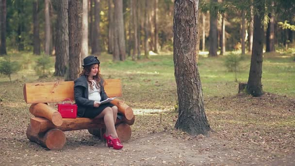 Kobieta z brązowymi włosami w kapeluszu w czerwonych butach i czerwoną torbą na ławce w parku jesienią czyta książkę. — Wideo stockowe