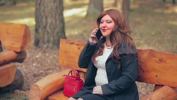 Μια γυναίκα με καστανά μαλλιά και μια κόκκινη τσάντα σε ένα παγκάκι το φθινόπωρο μιλάει τηλεφωνικώς — Αρχείο Βίντεο