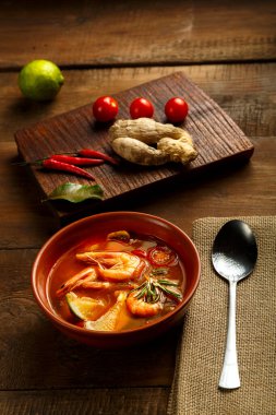 Tom yer elması çorbası, karides ve limonlu, kahverengi bir tabakta, ahşap bir masada, peçetedeki kaşığın yanında ve limonlu zencefil karışımı..