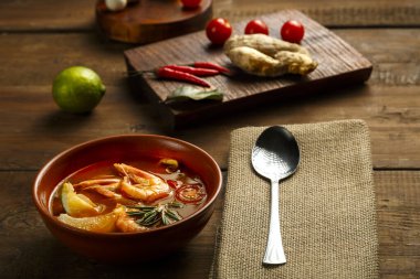 Tom yer elması çorbası, karides ve limonlu. Kahverengi bir tabakta, ahşap bir masanın üstünde. Peçetedeki kaşığın yanında..