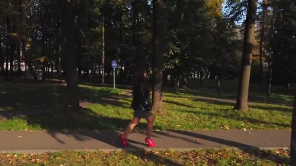 Женщина с каштановыми волосами в красных сапогах и красной сумкой гуляет в осеннем парке в солнечный день. — стоковое видео