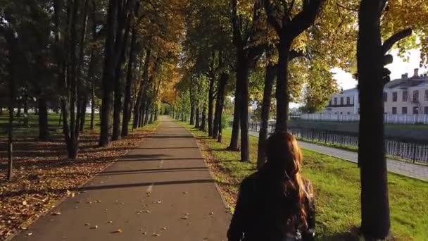 Nainen, jolla on ruskeat hiukset punaisissa saappaissa ja punainen pussi, ammutaan selästä, kun hän kävelee syksyn puistossa aurinkoisena päivänä. — kuvapankkivideo