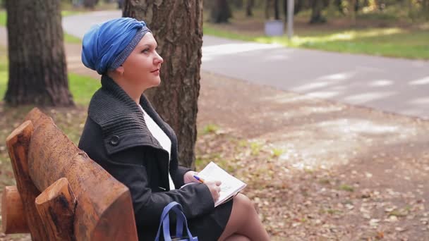 Eine Jüdin in traditioneller blauer Kopfbedeckung im Profil auf einer Bank in einer Gasse zeichnet in einem Notizbuch — Stockvideo