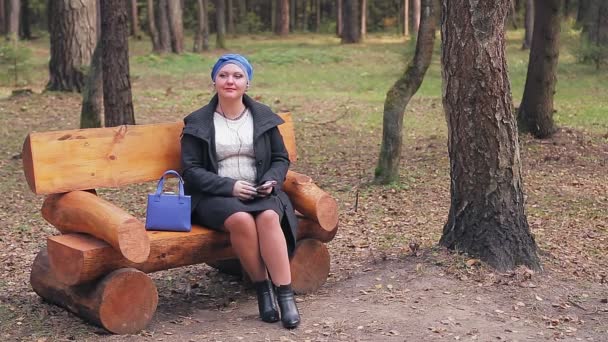 Mulher judia em um perfil de cobertura para a cabeça azul tradicional em um banco em um parque de outono escuta música com fones de ouvido — Vídeo de Stock