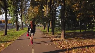 Arka manzara. Kahverengi saçlı, kırmızı çizmeli ve kırmızı çantalı bir kadın güneşli bir günde sonbaharda bir ara sokakta yürüyor..
