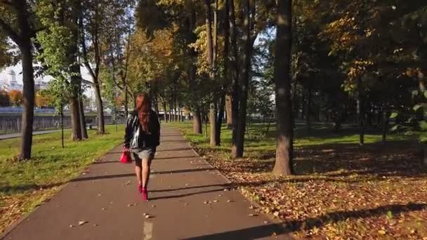 Hátsó nézet Egy barna hajú nő piros csizmában és piros táskával sétál egy sikátorban ősszel egy napsütéses napon. — Stock videók