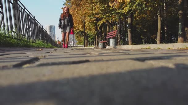 Наближення Жінка коричневого волосся в червоних чоботях і червона сумка прогулюється вздовж золотої осінньої алеї в сонячний день — стокове відео