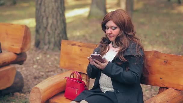 Eine Frau mit braunen Haaren und roter Tasche tippt im Herbst auf einer Parkbank in ein Smartphone. — Stockvideo