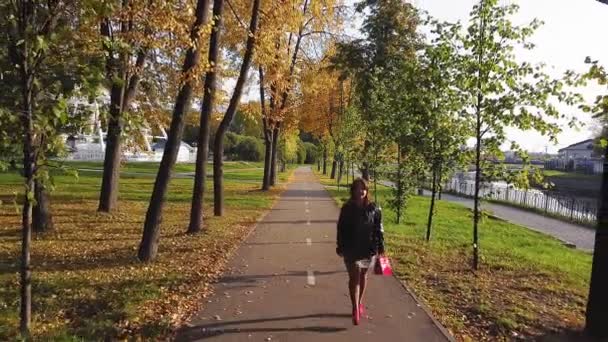 Vista trasera Una mujer de pelo castaño con botas rojas y una bolsa roja camina por un callejón en otoño en un día soleado — Vídeo de stock